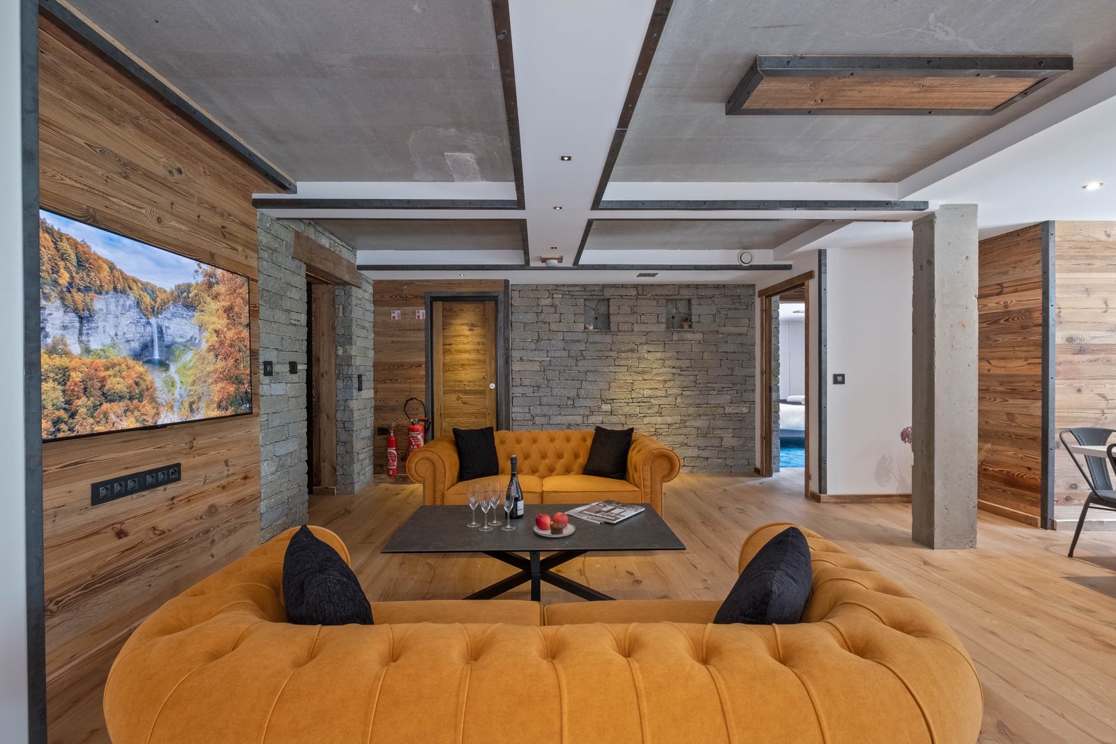 Solstice's living room in the OFF-PISTE Collection, Tignes, with polished concrete ceiling beams, a stone accent wall, and mustard chesterfield sofas