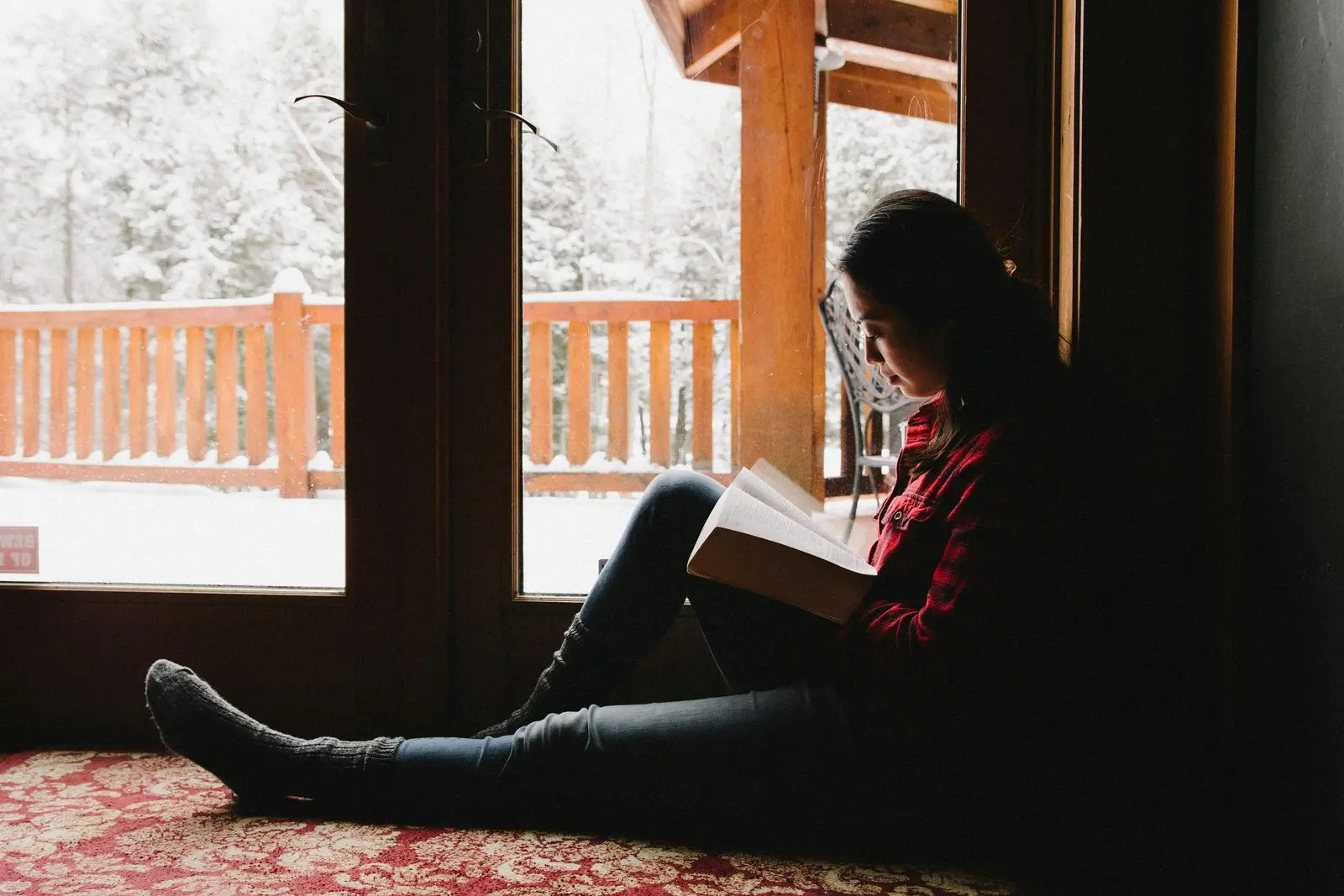 A woman reading a book on the floor by a chalet window, snow-covered forest outside