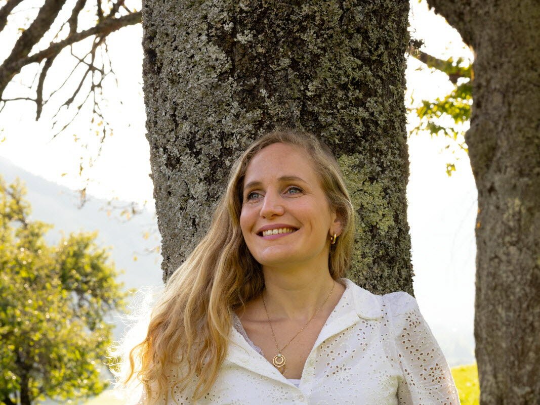 Novelist Sarah Séguigne smiling against a tree trunk, mountains in the background