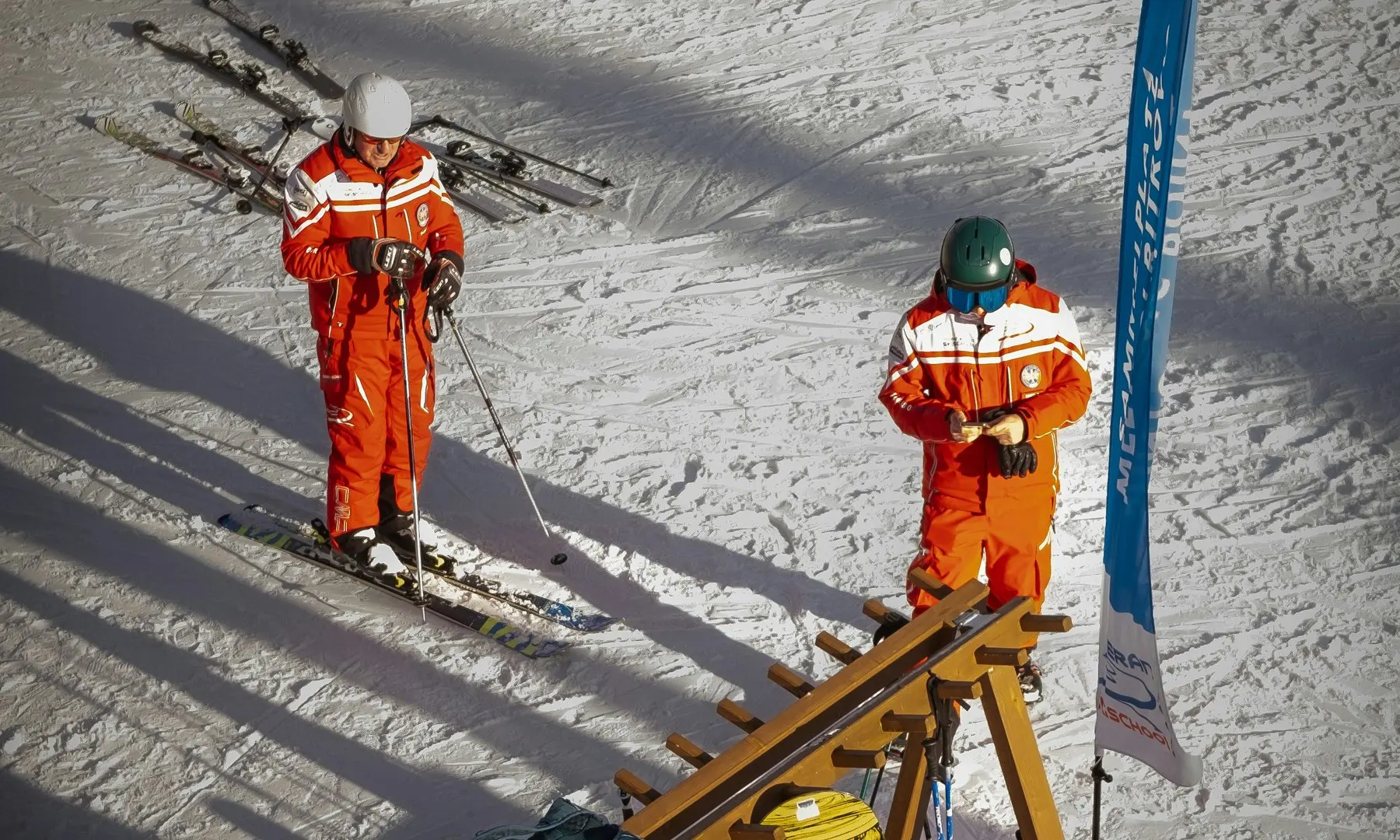Two ski instructors in orange uniforms preparing gear at a resort meeting point in the French Alps